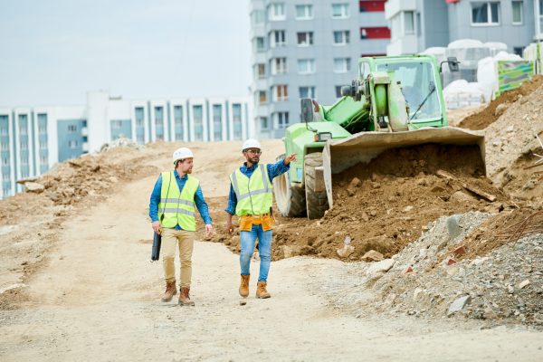 Two guys in helmets and waistcoats walking near digger and pointing at distance during inspection on construction site